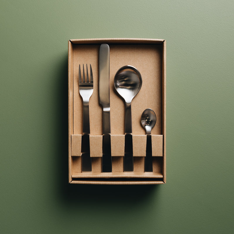 Acme's cutlery set in its cardboard, recyclable packaging. Flat lay image on a green background. Cutlery set contains Acme's fork, knife, spoon and teaspoon.